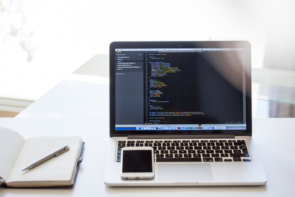 A silver laptop on a white desk displaying a code editor with lines of multicoloured programming code, positioned in front of a bright window with soft natural light. To the left of the laptop, there 