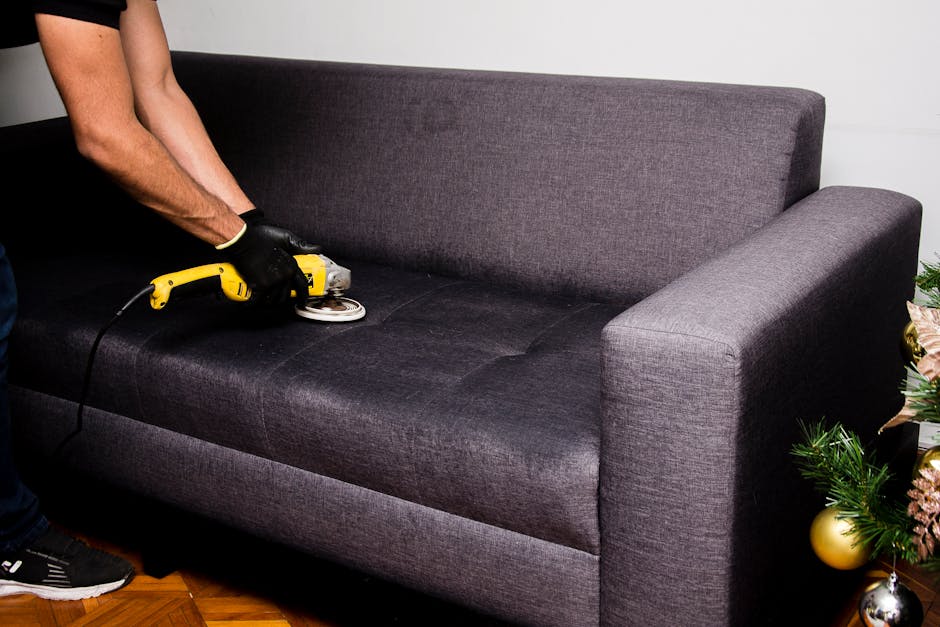 A woman vacuuming a black leather sofa in a bright, modern living room with large window panes, wooden ceiling panels, and contemporary decor. The room is well-lit with natural sunlight, showcasing a 