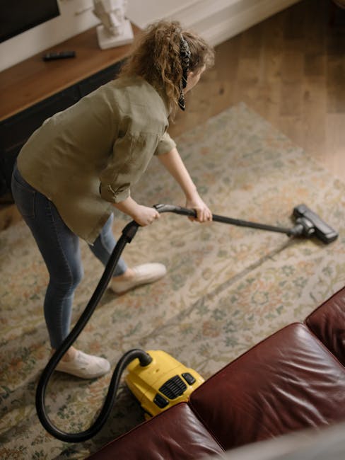 A person, wearing beige shoes and grey jeans, is operating a vacuum cleaner with a black hose on a patterned area rug featuring beige, green, and orange tones. The yellow vacuum cleaner is positioned 